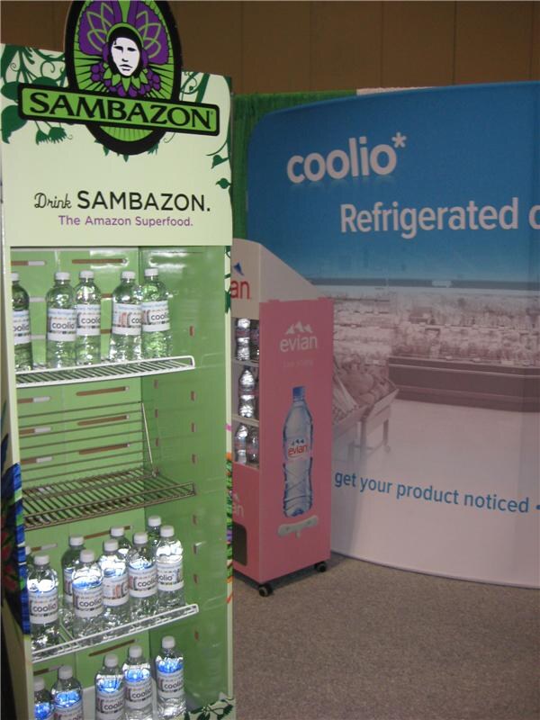 A green Sambazon display holds coolio-labeled water bottles beside a pink evian display. A blue coolio banner stands behind them.
