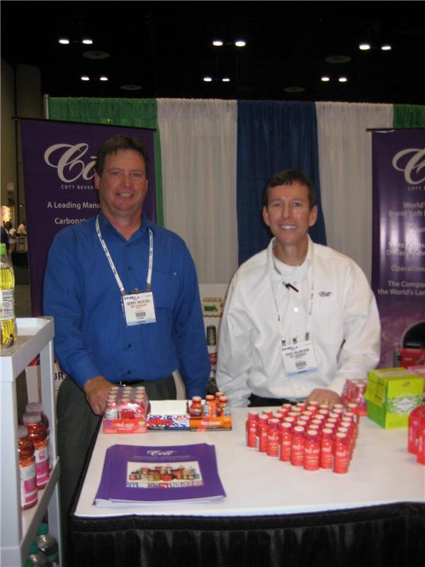 Two men with show badges stand behind a table of small red bottles and brochures. Purple Cott banners hang behind them.