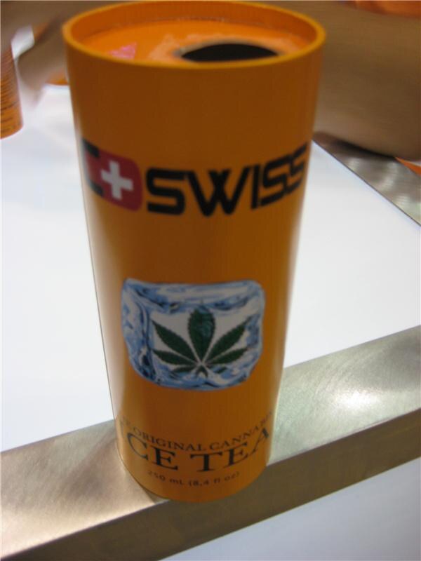 Close-up of an orange cylindrical C Swiss can of ice tea, 250 mL, with a green hemp leaf on an ice cube graphic, resting on a white and brushed-metal surface.
