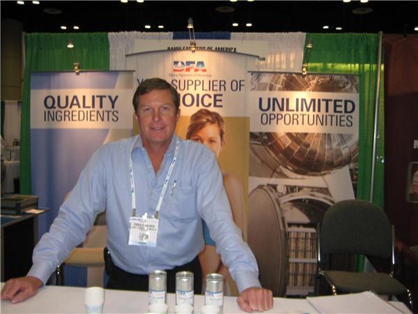 A man in a light blue shirt and lanyard stands behind a table with small canisters at the Dairy Farmers of America booth, with banners reading Quality Ingredients and Unlimited Opportunities.