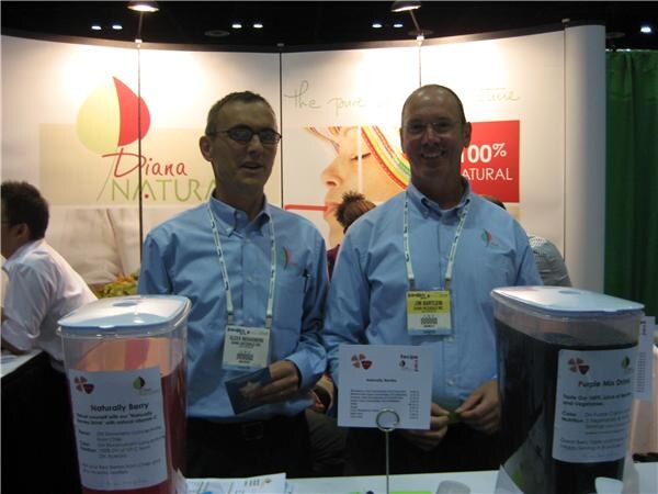 Two men in light blue shirts and lanyards stand at the Diana Naturals booth behind two large dispensers of red and dark purple drinks with printed labels.