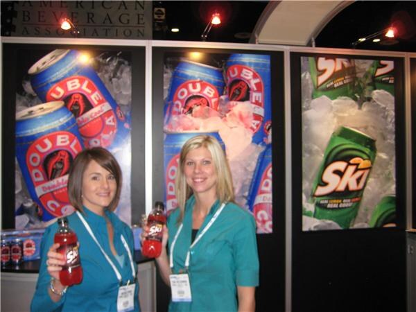 Two smiling women in teal shirts and lanyards hold bottles of Ski soda in front of large Double Cola and Ski can graphics at the booth.