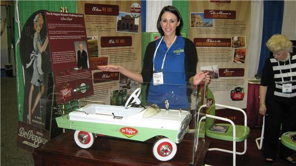 A woman in a blue top gestures toward a white and green Dr Pepper pedal car on display in the Dr Pepper Museum booth; an older woman stands at right.