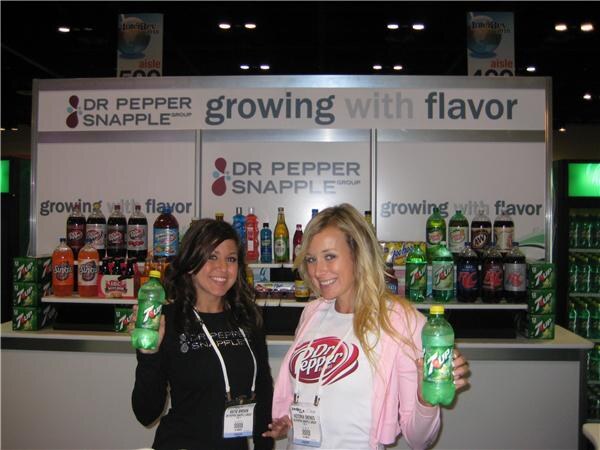 Two smiling women hold 7UP bottles at the Dr Pepper Snapple Group booth, with a Growing with Flavor sign and shelves of Dr Pepper and 7UP bottles behind them.