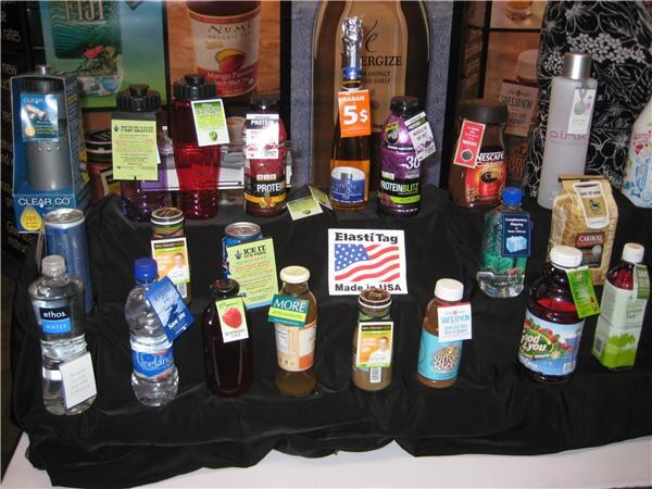 A black-draped display of bottles and cans, including Ethos Water and Nescafé, each with a small hanging tag, around a sign reading ElastiTag Made in USA.