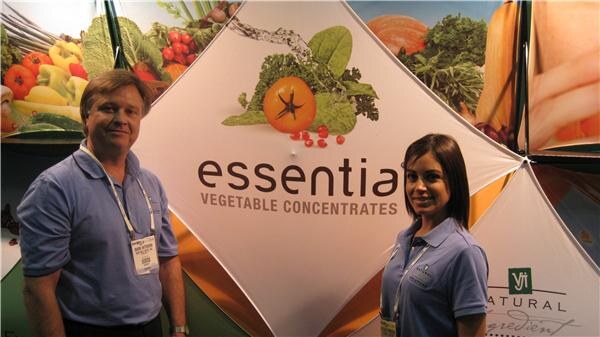 A man and a woman in blue polo shirts and badges stand in front of an essential Vegetable Concentrates display decorated with images of tomatoes and other vegetables.