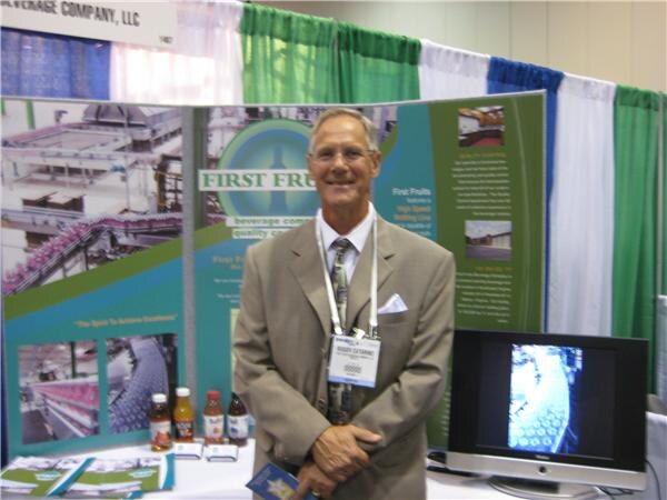 A smiling man in a tan suit and lanyard stands at the First Fruits booth, with display panels, a monitor and small bottles of tea on the table.