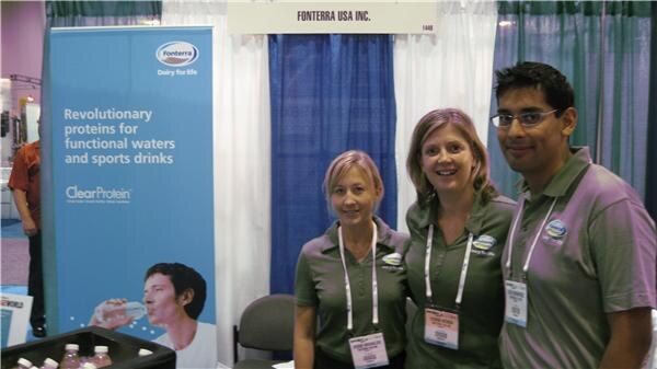 Two women and a man in gray-green polo shirts and lanyards stand at the Fonterra USA booth beside a ClearProtein banner.