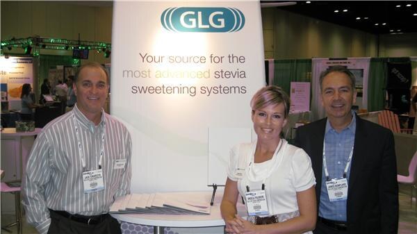 Two men and a woman with badges stand beside a GLG sign reading Your source for the most advanced stevia sweetening systems.