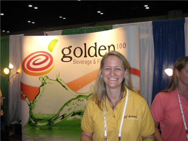 A smiling woman in a yellow polo and lanyard stands in front of a Golden 100 Beverage banner with a green splash graphic; another person in pink stands at right.
