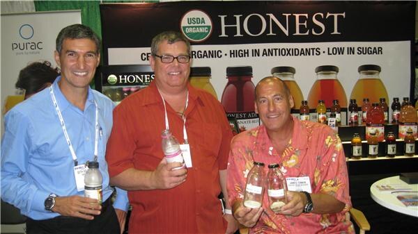 Three smiling men with badges stand in front of a Honest Tea booth backdrop with USDA Organic logo and bottles. Two hold bottles of tea. A Purac banner is at left.
