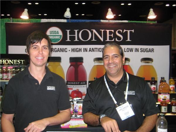 Two men in black Honest polo shirts stand smiling at the Honest booth, with bottles of Honest Ade and tea displayed on the counter and a USDA Organic banner behind them.