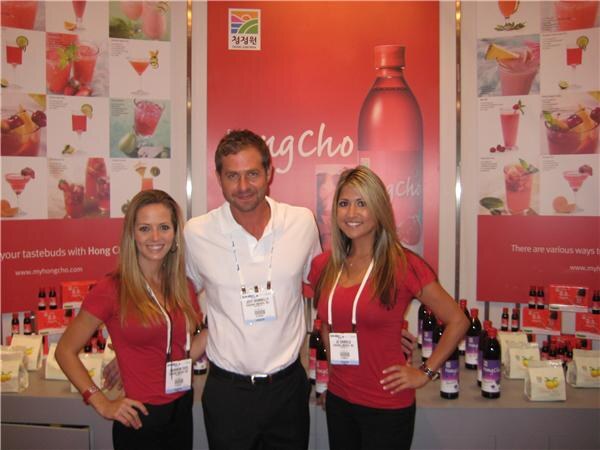 A man in a white polo and two women in red tops, all wearing badges, stand in the Hong Cho booth with a large red bottle graphic, cocktail images and bottles on the counter.