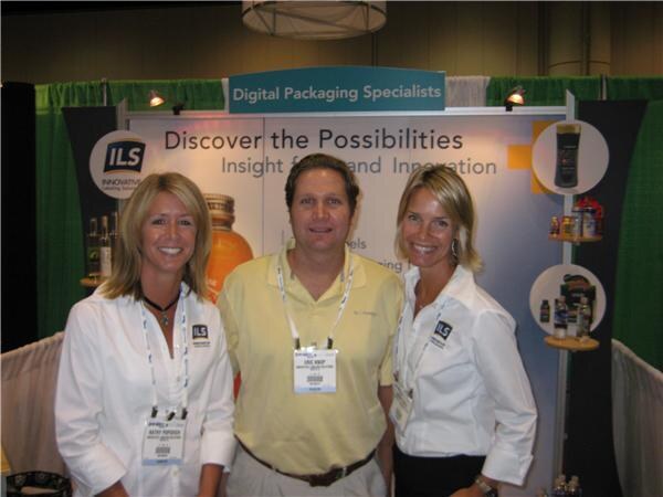 Two women in white shirts and a man in a yellow polo, all wearing badges, stand in the ILS booth under a Digital Packaging Specialists sign.