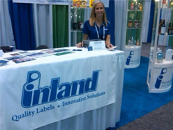 A woman in a blue polo stands behind a table draped with a white Inland cloth reading Quality Labels, Innovative Solutions. An IBWA member sign sits on the table, with display cases behind.