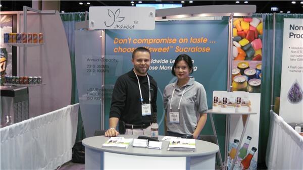 A man in a black shirt and a woman in a gray polo, both with badges, stand at a round table with brochures in the JKsweet booth, which has a sucralose banner behind them.