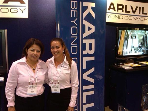 Two smiling women in pink shirts with badges stand in front of a blue Karlvil banner. A monitor and booth signage are visible beside them.