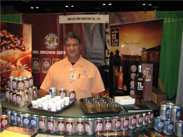 A man in an orange shirt stands behind a counter in the King Car Food Industrial booth, with Mr. Brown Coffee cans, sample cups, and a Kavalan whisky bottle and display.
