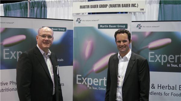 Two men in dark jackets stand in front of Martin Bauer Group banners in the company's booth under a sign naming Martin Bauer Group.