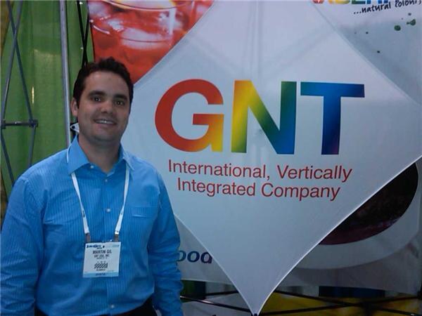 Martin Gil, in a blue striped shirt and lanyard, stands smiling in front of a GNT banner reading International, Vertically Integrated Company.
