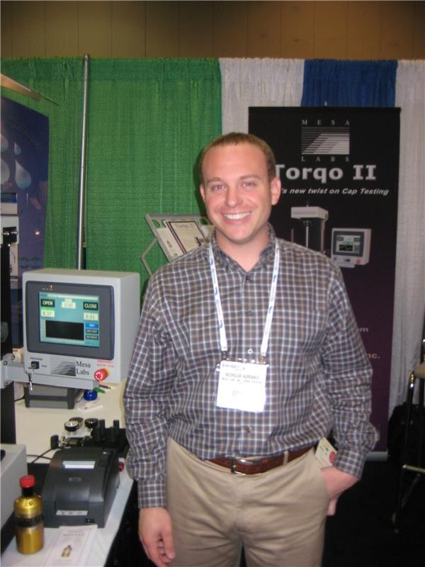 A smiling man in a plaid shirt and badge stands beside a Mesa Labs cap testing machine with a printer, in front of a Torqo II banner.