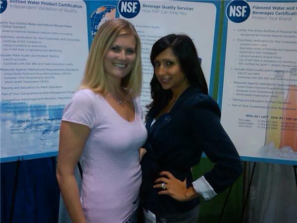 Two women pose in front of NSF posters about beverage quality services and bottled water certification. One has blonde hair and a pink top; the other has dark hair and a navy blazer.