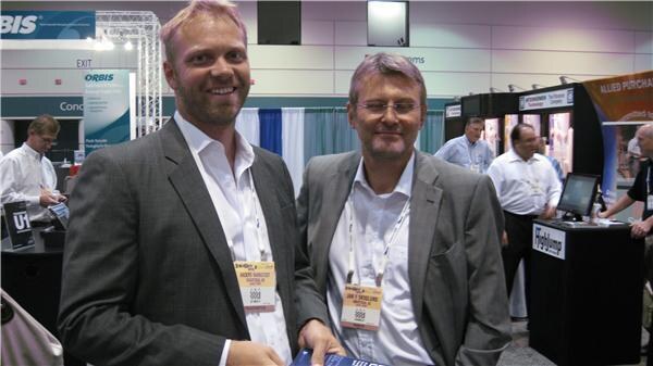 Two smiling men in gray jackets with badges stand in an aisle in front of ORBIS signage, with other attendees and booths in the background.
