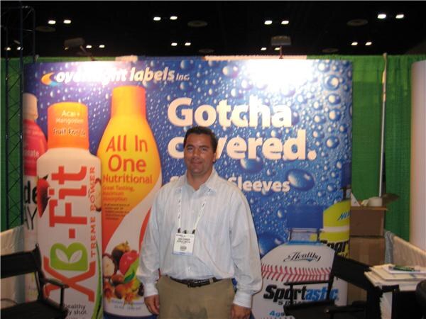 A man in a striped shirt with a badge stands in front of an Overnight Labels Inc. backdrop reading Gotcha covered, with large bottle graphics for X-Fit and Sportastic.