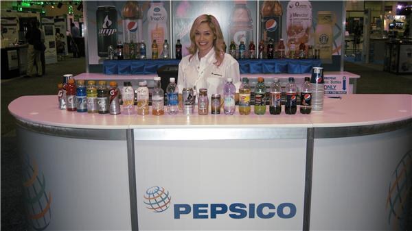 A smiling woman in a white jacket stands behind a pink PepsiCo counter lined with Pepsi, Gatorade and other bottled and canned drinks, with product banners including Amp behind her.