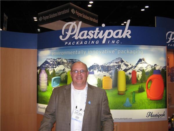 A bald man with glasses and a badge stands smiling in front of a Plastipak Packaging Inc. backdrop showing plastic bottles on a mountain landscape and the words environmentally innovative packaging.