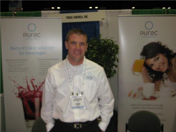 A man in a white shirt with a badge lanyard stands smiling between two Purac banners, one reading Nature's best solution for beverages and one showing a woman holding a glass of juice.
