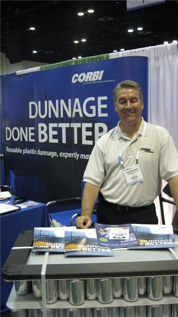 Richard Smith, in a cream Corbi Plastics polo, smiles and looks down at brochures on a cart. Behind him is a blue Corbi banner reading Dunnage Done Better.