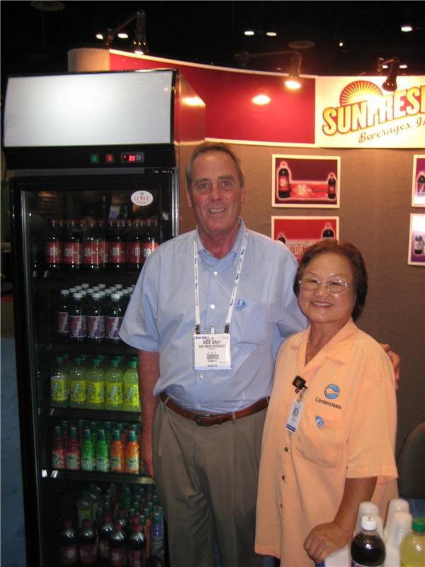 Rick Gray of Sun Fresh Beverages stands smiling beside a woman in a peach Centerplate shirt, in front of a lit refrigerator of bottled drinks and a Sunfresh Beverages sign.