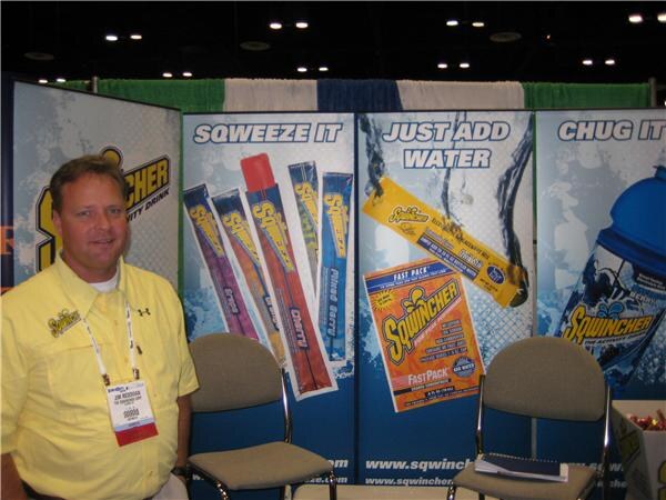 A man in a yellow Sqwincher shirt with a badge stands beside banners showing Sqwincher freezer pops, a Fast Pack powder packet and a bottle, with the phrases Squeeze It, Just Add Water and Chug It.