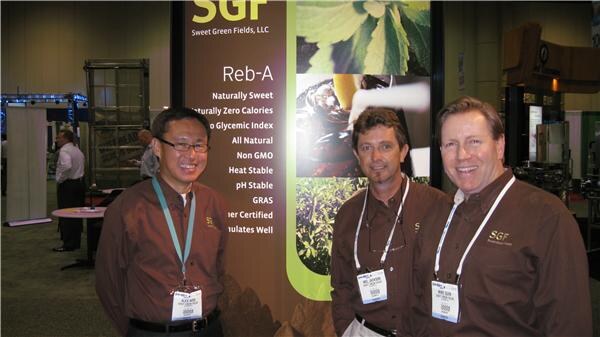 Three men in brown shirts with badges stand beside a Sweet Green Fields, LLC sign that lists Reb-A features such as naturally sweet, zero calories, non GMO and heat stable.