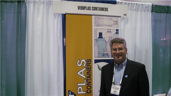 A smiling man with glasses, a dark blazer and a badge stands next to a yellow Veriplas Containers banner showing clear plastic bottles.