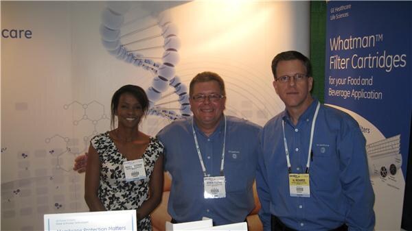 A woman and two men in blue shirts and badges stand together in front of a GE Healthcare Life Sciences display and a Whatman Filter Cartridges banner for food and beverage applications.