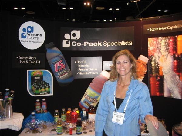 A smiling woman with long blonde hair and a name badge stands in the Winona Foods booth, with Co-Pack Specialists signage and bottled beverages on a table behind her.