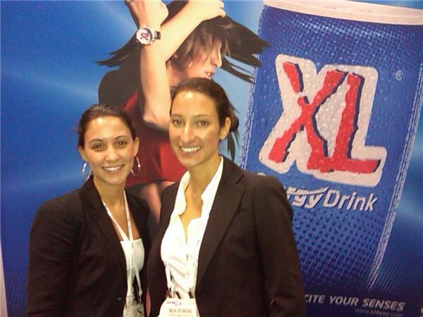 Two smiling women in dark blazers with lanyards stand in front of a large blue XL Energy Drink can graphic and a banner image of a woman.