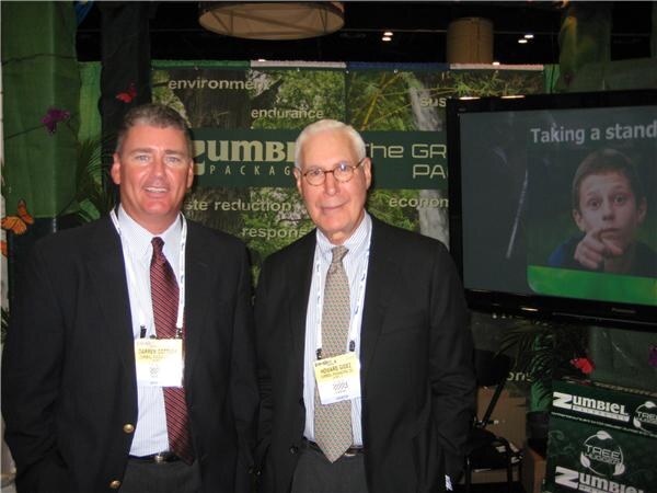 Two men in dark suits with badges stand side by side in the Zumbiel Packaging booth, with green signage and a screen showing a boy behind them.