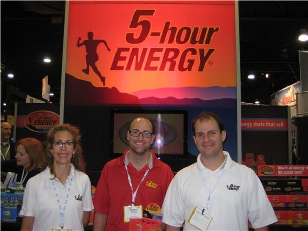 A woman and two men with badges stand under a large 5-hour ENERGY sign, with product boxes visible at right.