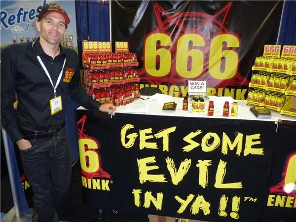 A smiling man in a black shirt and cap with a badge stands beside a 666 Energy Drink booth with bottles on shelves and a banner reading Get Some Evil In Ya.