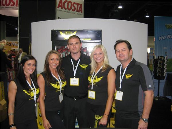 Five people in black ABB shirts with yellow wing logos stand together in front of a cooler, with an Acosta sign overhead.