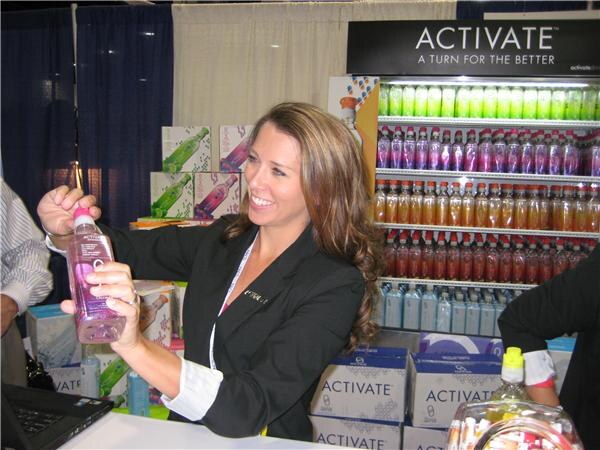 A smiling woman in a black blazer holds up a pink bottle at the Activate booth, with shelves of bottled drinks and Activate cartons behind her.