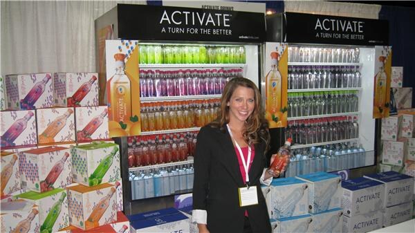 A smiling woman in a black blazer with a badge holds an orange Activate bottle in front of lit coolers and stacked Activate cartons.