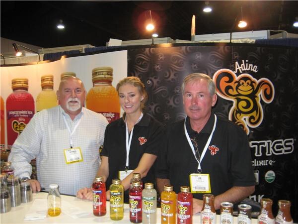 Two men and a woman in front of an Adina banner stand behind a counter holding bottled drinks, with large bottles on display behind them.