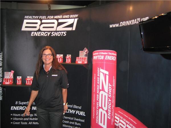 A smiling woman with glasses and a black polo stands in the Bazi booth, which has Bazi Energy Shots signs and a large pink Bazi shot display.