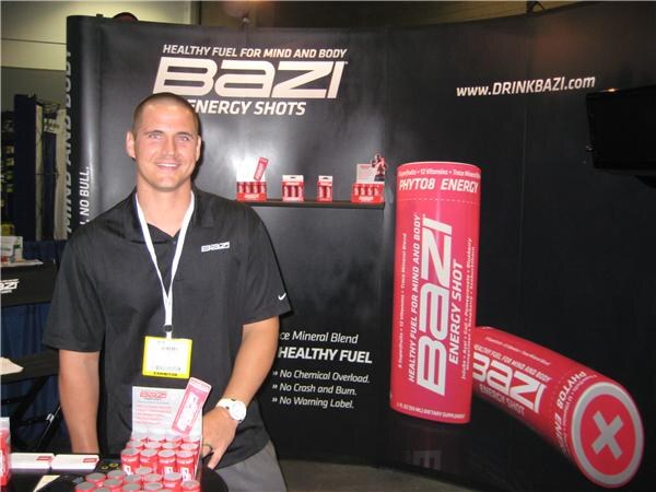 A smiling man in a black polo with a badge stands at the Bazi booth, with Bazi Energy Shots signage and large red shot props behind him.