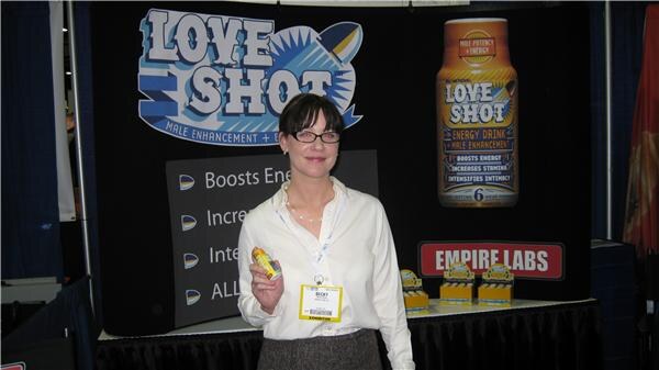 Becky, wearing glasses and a white blouse with a badge, holds a small bottle in front of a Love Shot backdrop and an Empire Labs sign.