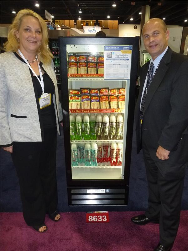 A woman in a light jacket and a man in a dark suit, both wearing badges, stand beside a Beverage-Air glass-door cooler stocked with wrapped sandwiches and bottled drinks.